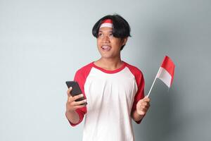 Portrait of attractive Asian man in t-shirt with red and white ribbon on head, holding mobile phone while raising up Indonesia flag. Isolated image on gray background photo