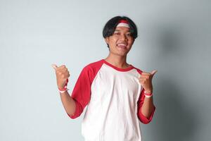 Portrait of attractive Asian man in t-shirt with red and white ribbon on head, showing good job hand gesture with thumbs up. Isolated image on gray background photo