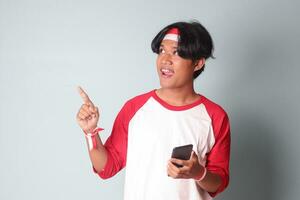 Portrait of attractive Asian man in t-shirt with red and white ribbon on head, holding mobile phone while raising up Indonesia flag and pointing away. Isolated image on gray background photo