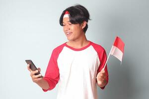 Portrait of attractive Asian man in t-shirt with red and white ribbon on head, holding mobile phone while raising up Indonesia flag. Isolated image on gray background photo