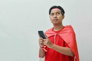 Portrait of attractive Asian man in t-shirt with red white ribbon on head with flag on his shoulder as a cloak, holding mobile phone and thinking looking up. Isolated image on gray background photo