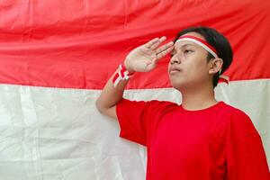 Portrait of attractive Asian man in red shirt with red and white ribbon on head, giving salute pose with hand in front of Indonesian flag photo