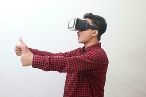 Portrait of Asian man in red plaid shirt using Virtual Reality VR glasses and showing thumb up hand gesture. Isolated image with copy space on white background. photo