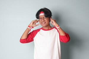 Portrait of attractive Asian man in t-shirt with red and white ribbon on head, having a migraine, touching his temple. Headache concept. Isolated image on gray background photo