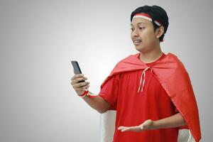 Portrait of attractive Asian man in t-shirt with red white ribbon on head with flag on his shoulder as a cloak, pointing and holding mobile phone. Isolated image on gray background photo