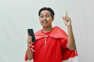 Portrait of attractive Asian man in t-shirt with red white ribbon on head with flag on his shoulder as a cloak, pointing and holding mobile phone. Isolated image on gray background photo