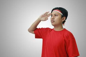 Portrait of attractive Asian man in t-shirt with red and white ribbon on head, giving salute pose with hand during Indonesian flag ceremony photo