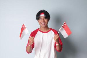 Portrait of attractive Asian man in t-shirt with red and white ribbon on head, showing product and poiting away while holding singapore flag. Isolated image on gray background photo