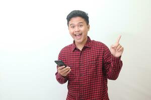 Portrait of attractive Asian man in casual plaid shirt standing against gray background, pointing away with his finger while using mobile phone. Advertising concept. photo