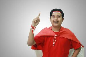 Portrait of attractive Asian man in t-shirt with red white ribbon on head with flag on his shoulder as a cloak, pointing up his forefinger and looking at camera. Isolated image on gray background photo