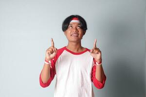 Portrait of attractive Asian man in t-shirt with red and white ribbon on head, pointing at something with finger. Isolated image on gray background photo