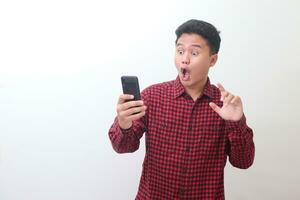 Portrait of shocked Asian man in casual plaid shirt standing against gray background, holding a mobile phone while trying to touch it. Advertising concept. photo