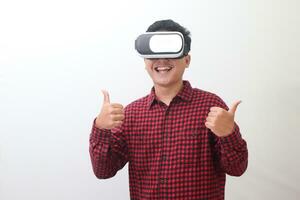 Portrait of Asian man in red plaid shirt using Virtual Reality VR glasses and showing thumb up hand gesture. Isolated image with copy space on white background. photo