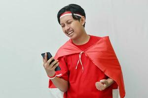 Portrait of attractive Asian man in t-shirt with red white ribbon on head with flag on his shoulder as a cloak, holding mobile phone with raised fist. Isolated image on gray background photo