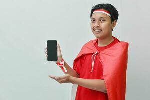 Portrait of attractive Asian man in t-shirt with red white ribbon on head with flag on his shoulder as a cloak, presenting a blank screen mobile phone. Isolated image on gray background photo