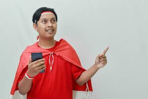 Portrait of attractive Asian man in t-shirt with red white ribbon on head with flag on his shoulder as a cloak, pointing and holding mobile phone. Isolated image on gray background photo