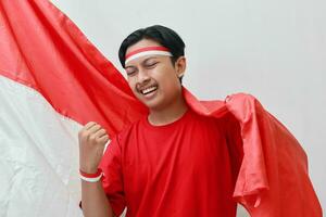 retrato de atractivo asiático hombre en camiseta con rojo y blanco cinta en cabeza, levantamiento bandera con su puño, celebrando de indonesia independencia día. aislado imagen en gris antecedentes foto