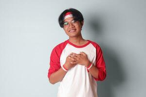 Portrait of attractive Asian man in t-shirt with red and white ribbon on head, placing hands on chest, feeling peaceful. Isolated image on gray background photo