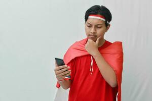 Portrait of attractive Asian man in t-shirt with red white ribbon on head with flag on his shoulder as a cloak, using mobile phone and holding hand on chin showing indecision. Isolated background photo