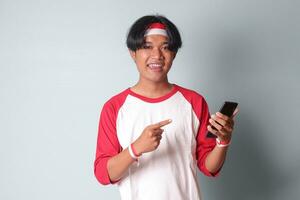 Portrait of attractive Asian man in t-shirt with red and white ribbon on head, holding mobile phone while raising up Indonesia flag and pointing away. Isolated image on gray background photo