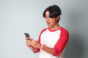retrato de conmocionado asiático hombre en camiseta con rojo y blanco cinta en cabeza, participación móvil teléfono con sorprendido expresión. aislado imagen en gris antecedentes foto