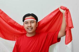 retrato de atractivo asiático hombre en camiseta con rojo y blanco cinta en cabeza, levantamiento bandera con su puño, celebrando de indonesia independencia día. aislado imagen en gris antecedentes foto