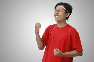 retrato de atractivo asiático hombre en camiseta con rojo y blanco cinta en cabeza, levantamiento su puño, celebrando de indonesia independencia día. aislado imagen en gris antecedentes foto