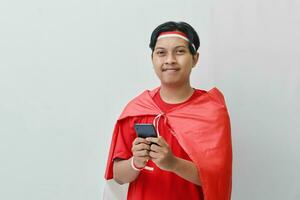Portrait of attractive Asian man in t-shirt with red white ribbon on head with flag on his shoulder as a cloak, holding mobile phone and looking at camera. Isolated image on gray background photo