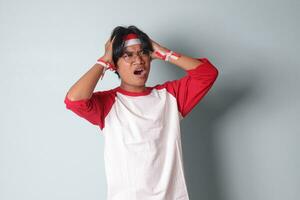 Portrait of attractive Asian man in t-shirt with red and white ribbon on head, having a migraine, touching his temple. Headache concept. Isolated image on gray background photo