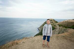 Little boy standing on top of a mountain and looking camera against lighthouse in sea. Cape Emine, Black sea coast, Bulgaria. photo