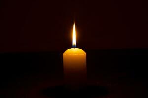 Single burning candle flame or light glowing on a big yellow candle on black or dark background on table in church for Christmas, funeral or memorial service with copy space. photo