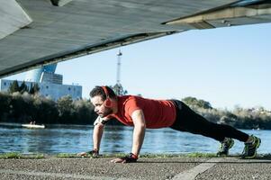 young man doing push-ups with a red t-shirt and music headphones, in an urban environment. healthy living concept photo