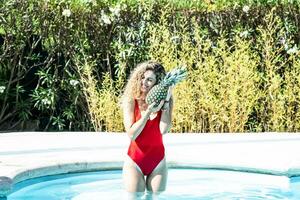 mujer sonriente en un rojo traje de baño dentro un piscina abrazando un piña foto