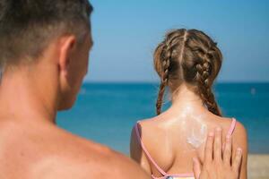 un padre aplica protector crema a su hija espalda a el playa. un hombre sostiene protector solar loción en un niño cuerpo. linda pequeño niña con protector solar por el mar. Copiar espacio. foto