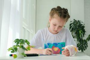 A little girl with a plaster on her arm is doing her homework. The child writes homework in a notebook. Hand injury. photo