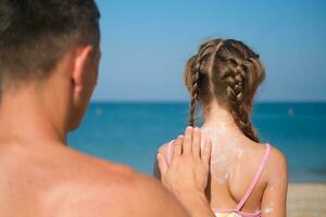 A father applies protective cream to his daughter's back at the beach. A man holds sunscreen lotion on a child's body. Cute little girl with sunscreen by the sea. Copy space. photo