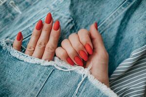 Beautiful female hands with a bright red matte manicure on a background of denim. Stylish oval nail design. Summer manicure. Copy space. photo