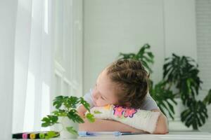 A little girl sleeps on a table with a broken arm. The child is bored doing homework, put her hand in plaster on the table. photo