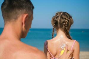 un padre aplica protector crema a su hija espalda a el playa. un hombre sostiene protector solar loción en un niño cuerpo. linda pequeño niña con protector solar por el mar. Copiar espacio. foto