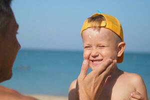 un padre aplica protector crema a su hijos cara a el playa. un del hombre mano aplica protector solar loción en un niño rostro. foto