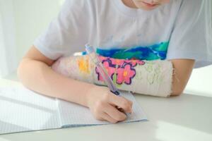 A little girl with a plaster on her arm is doing her homework. The child writes homework in a notebook. Hand injury. photo
