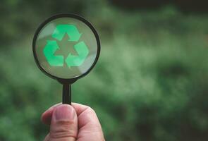 Concepts of waste reduction, pollution, reuse, efficient use of resources. Protect the environment by recycling. Human hand holding a magnifying glass with recycling sign on nature background. photo