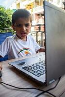 pequeño chico sentado a mesa utilizando ordenador portátil para en línea clase en grado 1, niño estudiando en ordenador portátil desde hogar para distancia aprendizaje en línea educación, colegio chico niños estilo de vida concepto foto