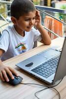 pequeño chico sentado a mesa utilizando ordenador portátil para en línea clase en grado 1, niño estudiando en ordenador portátil desde hogar para distancia aprendizaje en línea educación, colegio chico niños estilo de vida concepto foto