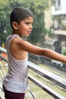 pequeño niño jugando en verano lluvia en casa balcón, indio inteligente chico jugando con lluvia gotas durante monzón lluvioso estación, niño jugando en lluvia foto
