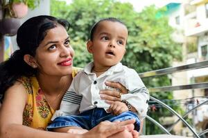 amoroso mamá que lleva de su bebé a hogar balcón. brillante retrato de contento mamá participación niño en su manos. madre con su pequeño 1 año antiguo hijo. foto