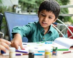 Smart Indian little boy perform thumb painting with different colourful water colour kit during the summer vacations, Cute Indian Kid doing colourful thumb painting drawing on wooden table photo