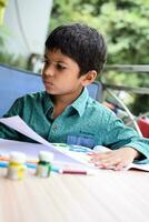 Smart Indian little boy perform thumb painting with different colourful water colour kit during the summer vacations, Cute Indian Kid doing colourful thumb painting drawing on wooden table photo