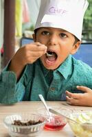 linda indio cocinero chico preparando helado con frutas y nueces plato como un parte de no fuego Cocinando cuales incluye vainilla hielo crema, duende, Coco polvo, recién Cortado frutas y fresa jarabe. pequeño niño preparando comida foto