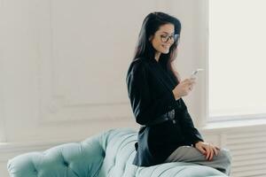 European entrepreneur in corporate suit and spectacles, using online banking app for money transfer, holding cellphone, sending emails, posing at the back of a comfortable sofa, utilizing free wifi. photo